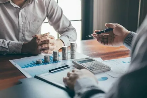 A financial advisor pointing a pen toward growth charts and coin stacks during a personal retirement planning consultation with a client
